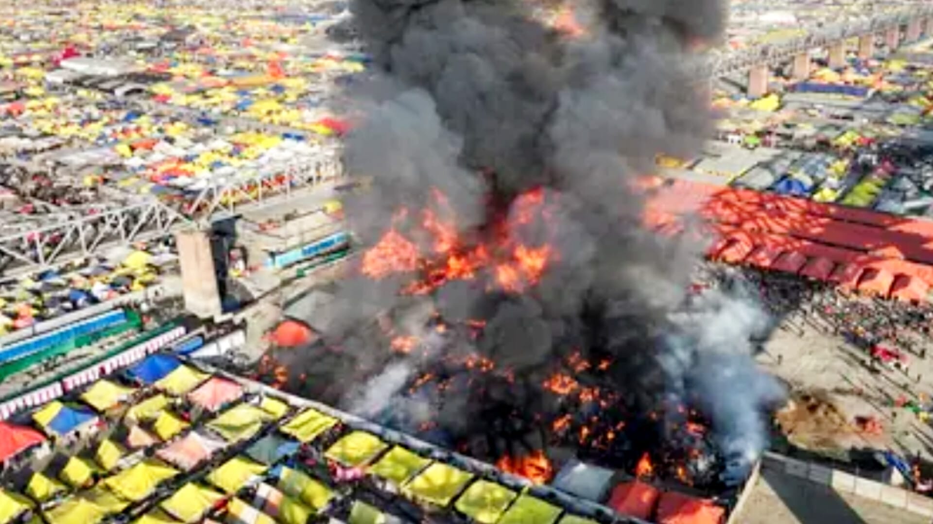 Aerial view of a massive fire in the Mahakumbh tent city in Prayagraj, with thick black smoke and flames engulfing several tents, surrounded by colorful tent structures and a large crowd.